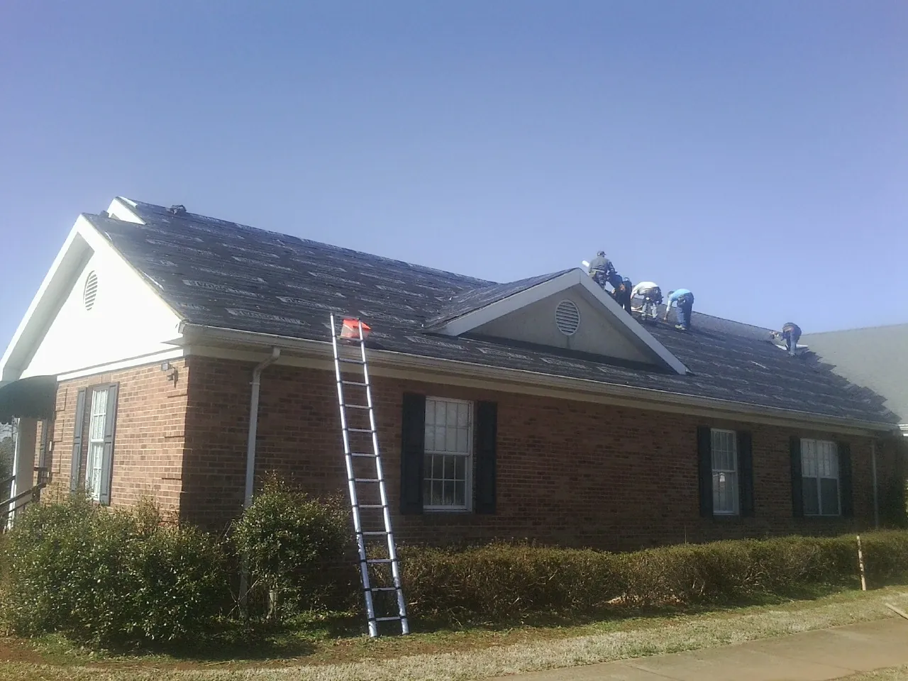 Crew tearing shingles off a brick ranch roof with a ladder set — Emergency Roof Repair work by our crew in Roundhill