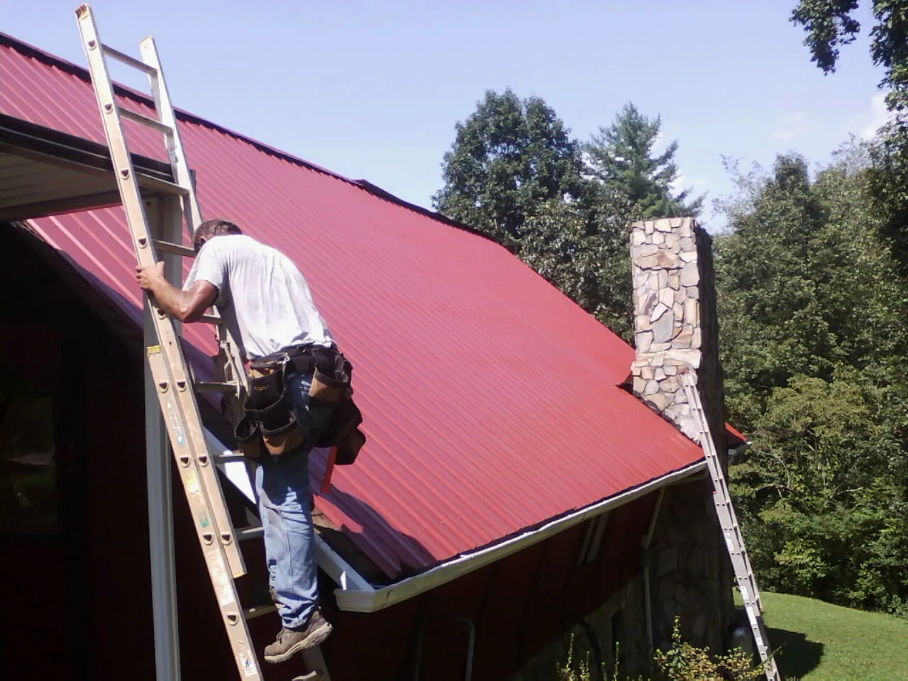 Roofer on a ladder beside a red metal roof and stone chimney — Soffit & Fascia Repair by Roofing Brothers in Roundhill